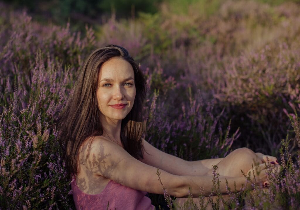 Anna sitzt auf einer Wiese und schaut seitlich zur Kamera. Sie hat braune lange Haare und lächelt leicht. Es ist Sommer. Um sie herum leuchten lila Blumen und grünes Gras.